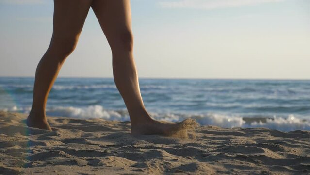 Female feet walking barefoot on the shore at sunny day. Legs of young girl going along sea beach. Seaside landscape at background. Summer vacation or holiday concept. Close up Slow motion
