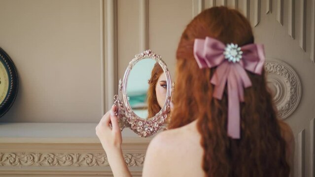 Close-up portrait red-haired girl fantasy princess looks at reflection in vintage silver mirror curly long hair beauty face. Woman sexy fashion model. Queen in red dress back rear view. classic room