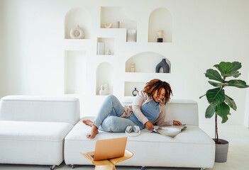 oung african woman sitting on sofa indoors at home while reading magazine or book