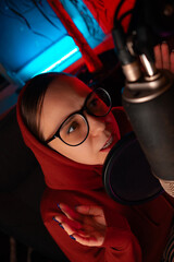 Portrait of a womam in a red hoodie and glasses recording a podcast live into a microphone.