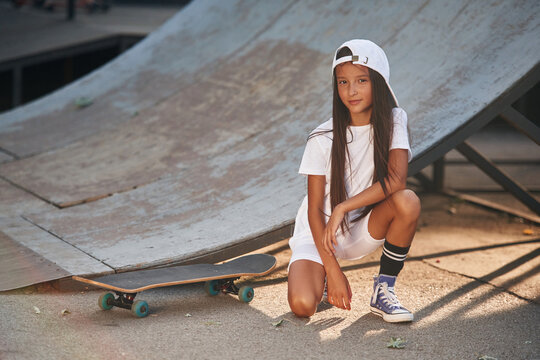 Ramp is behind. Girl is sitting. With skateboard outdoors