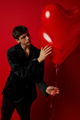 Obraz na płótnie Canvas romantic young man in velvet attire holding heart-shaped balloons on red background, Valentines day
