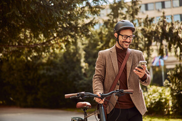 A grinning young tourist man checking a map on his phone and pushing a bicycle through a park.