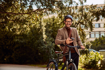 Fototapeta na wymiar Portrait of a smiling businessman holding a mobile phone and leaning a bicycle on a hip in a park.