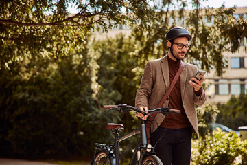 A businessman with a helmet on his head using a phone and pushing a bicycle.