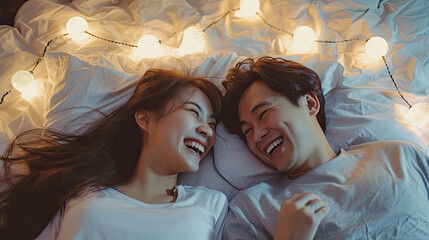 Lovely cute smiling young Asian lover couple lying on elbow on the bed and laughing while talking funny in the bedroom at home. Concept of romantic relationship.