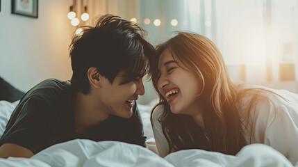 Lovely cute smiling young Asian lover couple lying on elbow on the bed and laughing while talking funny in the bedroom at home. Concept of romantic relationship. - obrazy, fototapety, plakaty