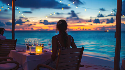 Romantic dinner on sunset. Woman sitting alone on table set with lantern for a romantic meal on beach, yachts and ocean on background. Dinner for a couple in love in luxury outdoor restaurant - obrazy, fototapety, plakaty
