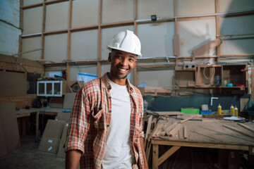 Happy African American male with hardhat stands in carpentry factory