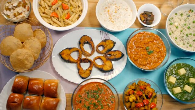 Variety of vegetarian and non-vegetarian food served on the dining table. Pan shot of selective dishes of Poori  Pulao  Pasta  Fish Fry  Pav Bhaji  Jeera-Aloo  and pickles spread on a wooden table