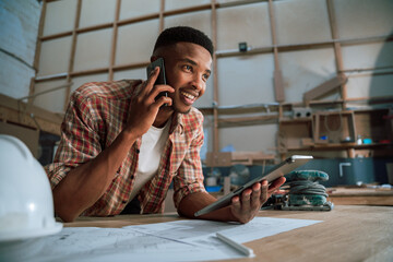 Smiling young African male uses cellphone while holding tablet in woodwork warehouse 