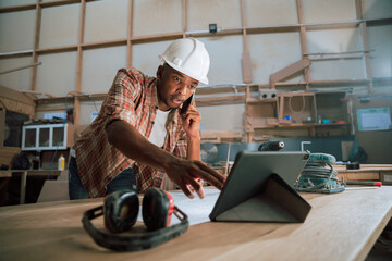 Concentrated African male uses tablet while on the phone in a woodwork factory  - obrazy, fototapety, plakaty