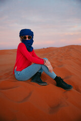 Young girl sitting on top of a dune in the sahara at sunset with a berber scarf