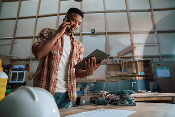 Young African male in a carpentry factory speaks on the phone while holding tablet