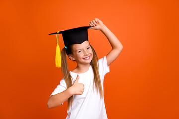 Portrait of clever funny small girl hand touch mortarboard hat demonstrate thumb up isolated on orange color background