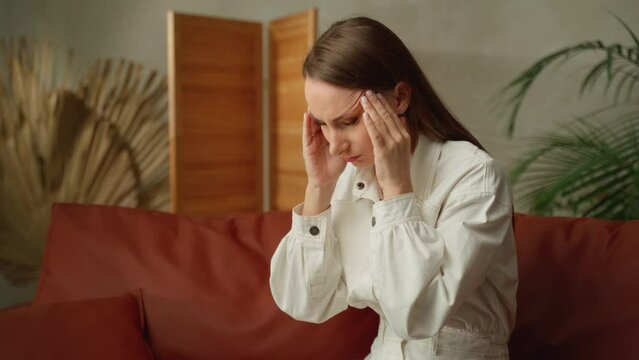 A tired woman with a severe headache. A young girl in a state of stress, suffering from acute migraine or pain with temporal arteritis, is sitting at home on the couch. 