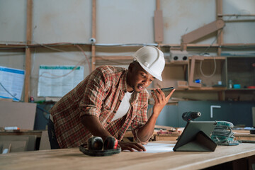 Young African male looks at designs while using tablet and phone in woodwork factory 