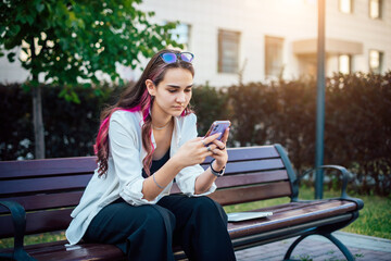 Obraz premium Portrait shot of a young brunette woman messaging on mobile phone while sitting on the bench outdoors. Beautiful female person using smartphone outside.