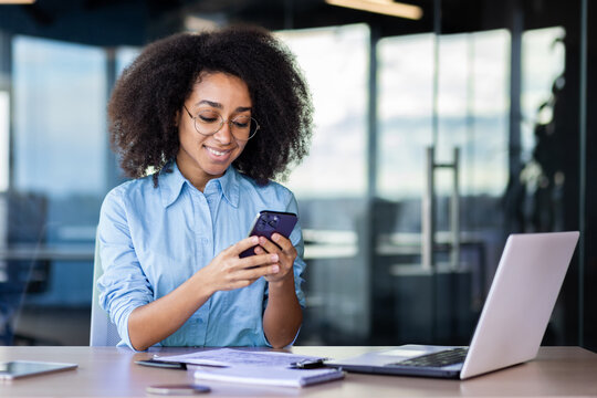 Businesswoman at workplace inside office, employee uses app on phone, woman happily smiles browsing social media and reading text message.