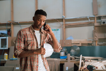 Young African male holds hardhat while on the phone in carpentry warehouse 