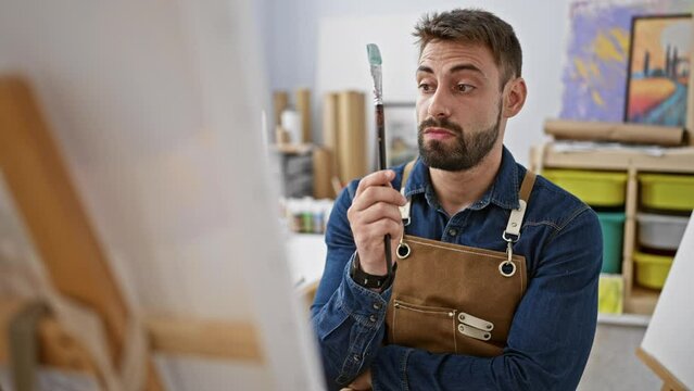 Focused young hispanic man artist seriously contemplating his painting at an art studio, doubt lingering as he draws but driven by a burning idea