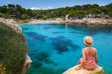 Fototapeta na wymiar A woman leisurely sitting on a cliff, looking out at Cala Gat beach in Mallorca