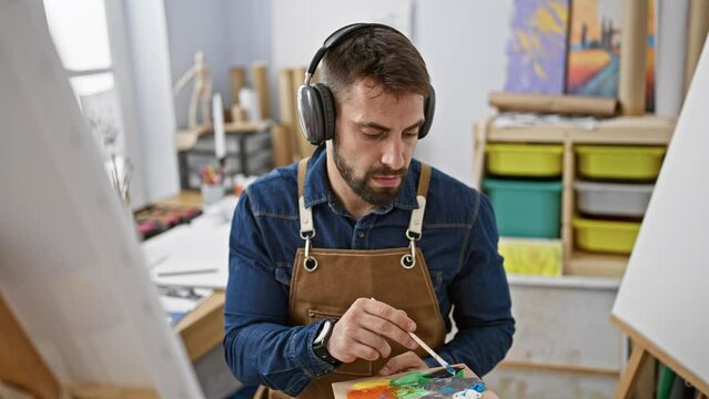 Young hispanic male artist, seriously focused at the art studio, drawing a portrait, while listening to music amidst brushes, canvas, and paint