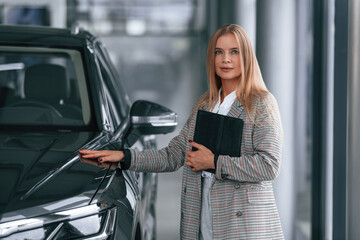 Fototapeta premium With tablet in hands. Woman in formal clothes is in the car dealership