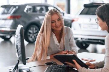 Sitting by the table. Female manager is helping woman customer in the car dealership salon