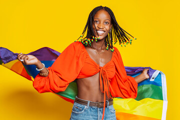 African american young smiling girl with braids, toothy smiles with pride rainbow flag in motion at studio isolated over yellow background. Trans or bisexual concept. - obrazy, fototapety, plakaty