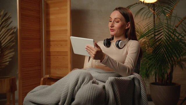 Video conference. Portrait of a cheerful woman in a headset making a web call using a tablet computer, sitting on an upholstered chair, talking, waving at a webcam. 
