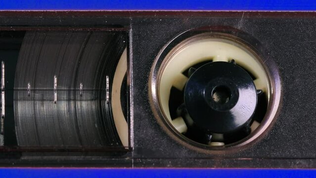 Nostalgic audio cassette tape unwinds behind a transparent window in a blue plastic cover, the tapes reels, percent marks, and other details coming into view as the camera pans left. Extreme Close up
