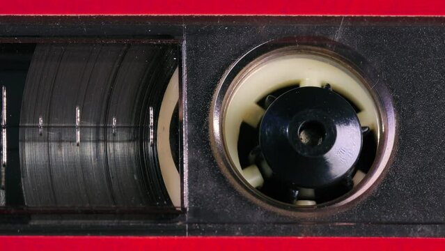 Nostalgic audio cassette tape unwinds behind a transparent window in a red plastic cover, the tapes reels, percent marks, and other details coming into view as the camera pans left. Extreme Close up
