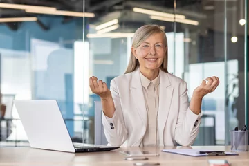  Portrait of senior mature business woman boss at workplace inside office, woman smiling and looking at camera resting meditating in lotus position, practicing yoga, working with laptop. © Liubomir