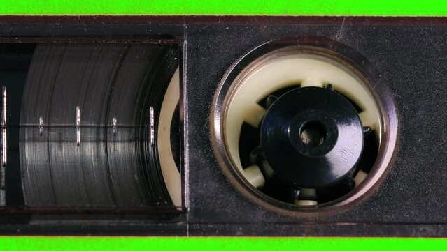 Nostalgic audio cassette tape unwinds behind a transparent window in a green plastic cover, the tapes reels, percent marks, and other details coming into view as the camera pans left. Extreme Close up