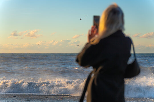Young woman in a black coat photographs huge waves in the sea, seagulls at sunset, focus on the sea. Stormy sea and a woman with a smartphone.