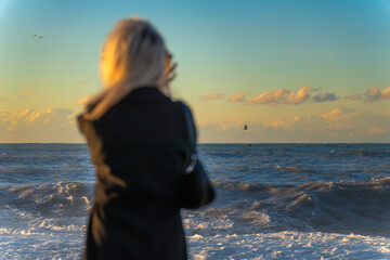 Blonde young woman in a black coat looks at a stormy sea or ocean at sunset, a selective focus. Photo from the back of a woman looking over the horizon of the sea