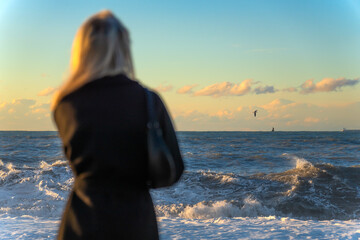 Blonde young woman in a black coat looks at a stormy sea or ocean at sunset, a selective focus. Photo from the back of a woman looking over the horizon of the sea