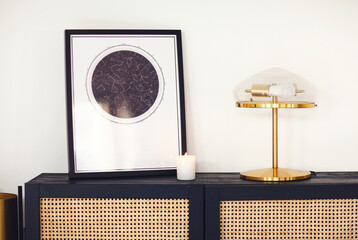 Black wooden commode with framed star map poster, lamp and candle in minimalist room interior