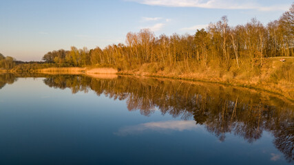 Fototapeta na wymiar This serene image captures the calm waters of a river during the golden hour, with the warm light of the setting sun illuminating the trees and grasses along the banks. The trees, stripped of their