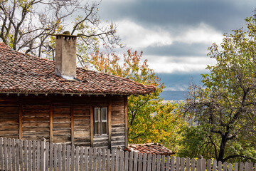 Fototapeta na wymiar Old time traditional rural house with wooden walls and windows, fence. Autumn yellow leaves of trees, roof with clay tiles and chimney. Typical architecture from the Bulgarian National Revival period.
