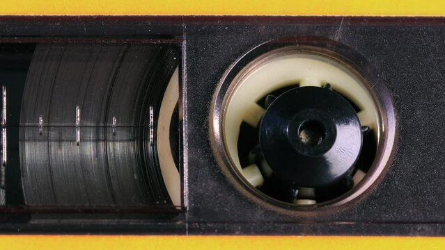 Nostalgic audio cassette tape unwinds behind transparent window in a yellow plastic cover, the tapes reels, percent marks, and other details coming into view as the camera pans left. Extreme Close up
