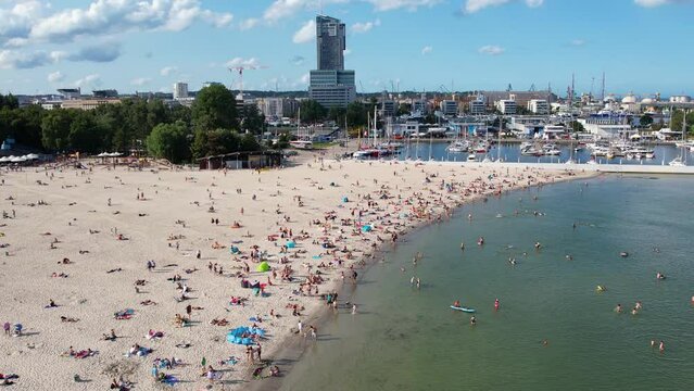 Beach in a modern seaside city Gdynia, aerial view. Establishing shot of crowded beach near yacht port and marina. Sunbathing by the Baltic sea in a sunny summer day.