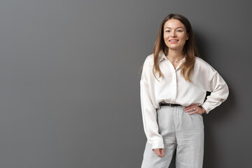 A relaxed and confident professional woman in a white blouse and grey trousers, exuding casual elegance against a grey background