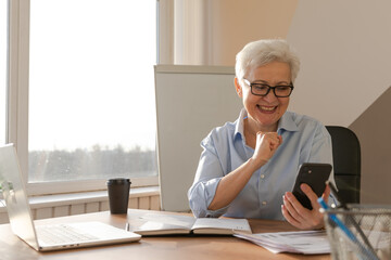 Confident stylish european middle aged senior woman using smartphone at workplace. Stylish older...
