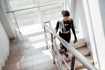 Cheerful sporty brunette woman with drink walking on stairs in gym in sunny day.