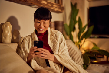 A smiling Asian woman using a mobile phone while resting on the couch.