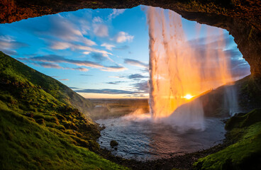 Impressive waterfall “Seljalandsfoss“ in Iceland seen from a cave behind the sunlit water curtain at midnight in June. Colorful sunset panorama in wild nature of volcanic scenery. Major attraction.