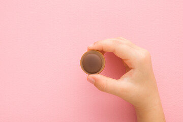 Little child fingers holding brown caramel chocolate candy on light pink table background. Pastel color. Sweet snack. Closeup.