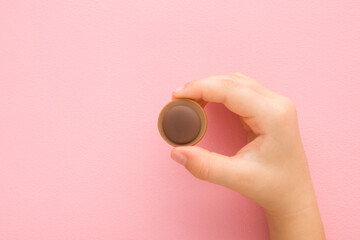 Little child fingers holding brown caramel chocolate candy on light pink table background. Pastel...